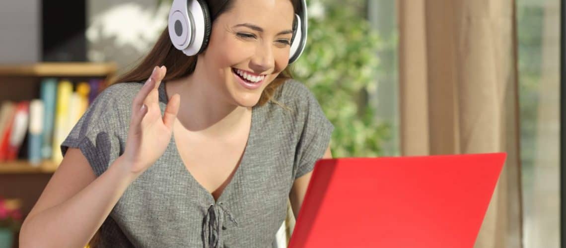 Happy woman wearing headphones greeting during a video conference on line with a red laptop sitting in a table in the living room